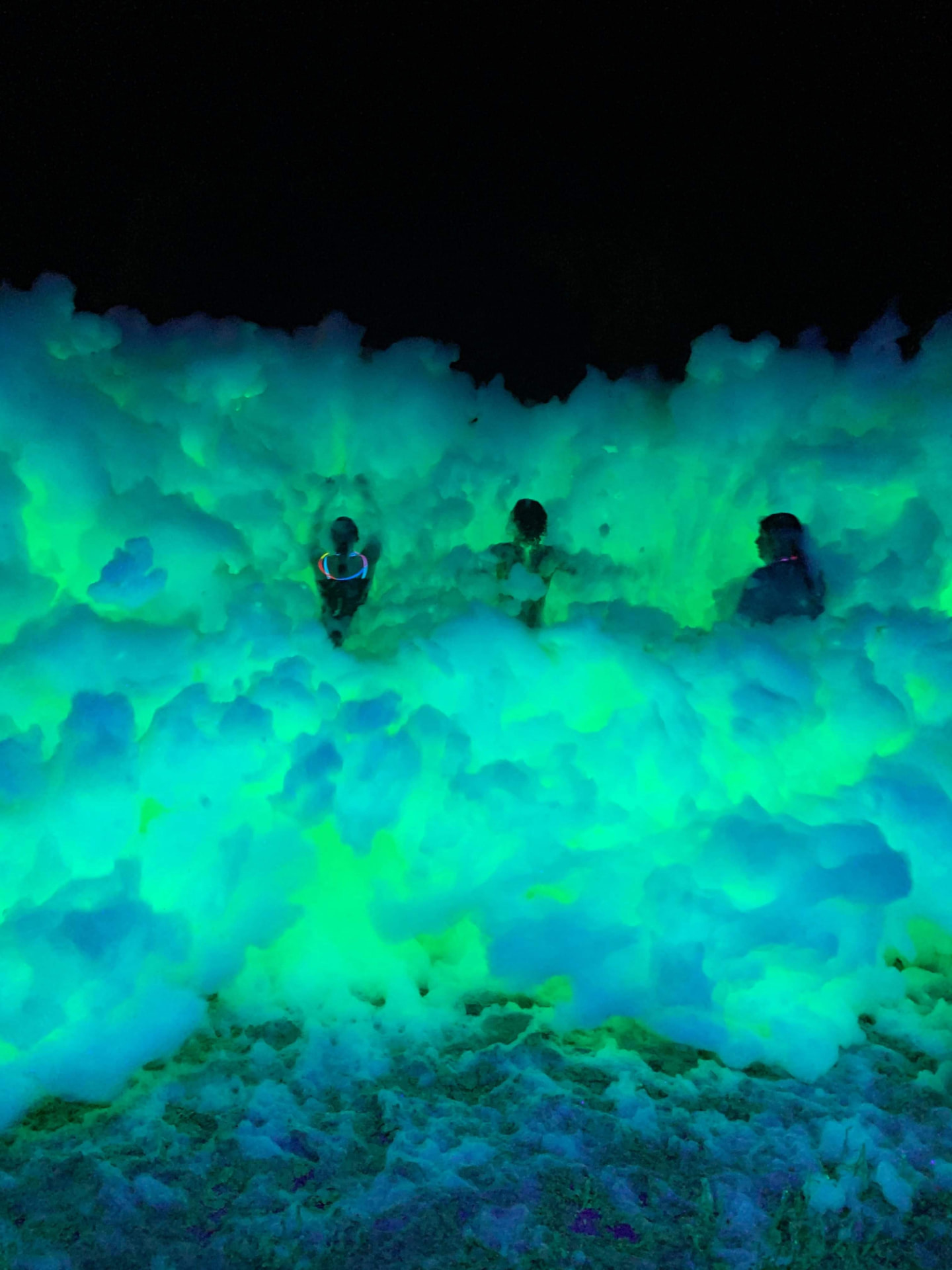 Kids surrounded by a huge wave of glowing green and blue UV foam at night